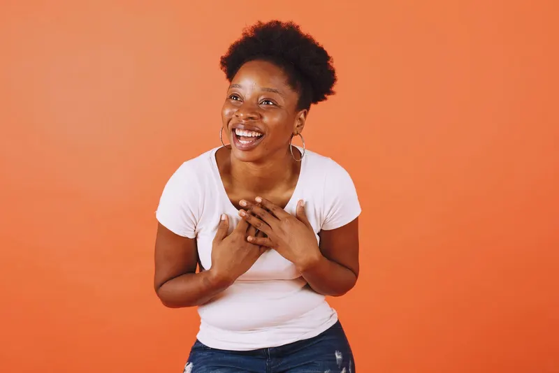A woman laughing with her hands on her chest against an orange backdrop