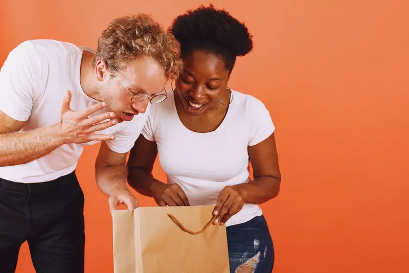 Two friends laughing over a paper bag on an orange backdrop